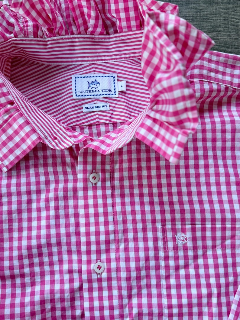 May include: A pink and white gingham-patterned shirt with a ruffled collar. The shirt features a button-down front, a chest pocket with a small embroidered fish, and a label that reads "SOUTHERN TIDE CLASSIC FIT".