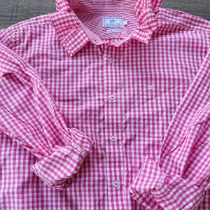 May include: A pink and white gingham button-down shirt with long sleeves and a ruffled collar. The shirt features a classic fit and a small logo on the chest. The sleeves are rolled up at the cuffs.
