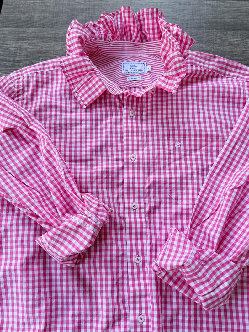 May include: A pink and white gingham button-down shirt with long sleeves and a ruffled collar. The shirt features a classic fit and a small logo on the chest. The sleeves are rolled up at the cuffs.