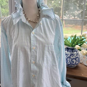 May include: A light blue and white checkered shirt with a ruffled collar and a pearl necklace. The shirt is buttoned up and has a pocket on the chest. The shirt is displayed on a mannequin.