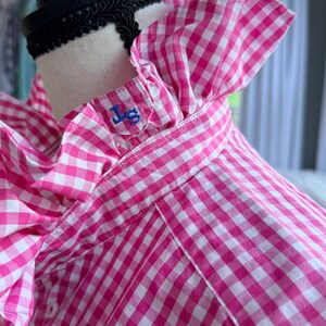 May include: A pink and white gingham shirt with a ruffled collar and sleeves. The shirt has a blue embroidered monogram on the collar. The shirt is displayed on a mannequin.