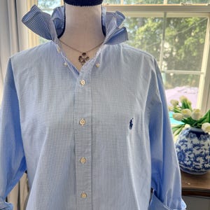 May include: A light blue and white gingham button-down shirt with a high collar, long sleeves with rolled cuffs, and a small dark blue embroidered logo on the left chest. The shirt is displayed on a mannequin with a black choker and a silver necklace.