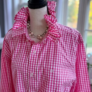May include: A pink and white gingham shirt with a ruffled collar and cuffs. The long-sleeved shirt has a button-down front and a small pocket on the chest. A pearl necklace is draped around the collar.