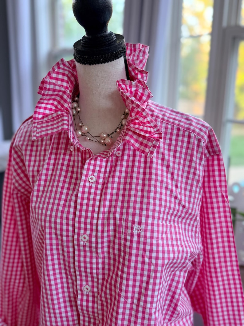 May include: A pink and white gingham shirt with a ruffled collar and cuffs. The long-sleeved shirt has a button-down front and a small pocket on the chest. A pearl necklace is draped around the collar.