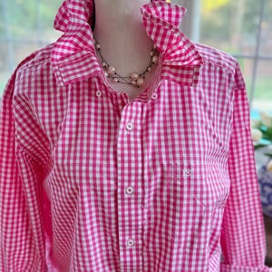 May include: A pink and white gingham button-down shirt with a ruffled collar and cuffs. The long-sleeved shirt has a small pocket on the chest and is displayed on a mannequin. A pearl necklace is visible.