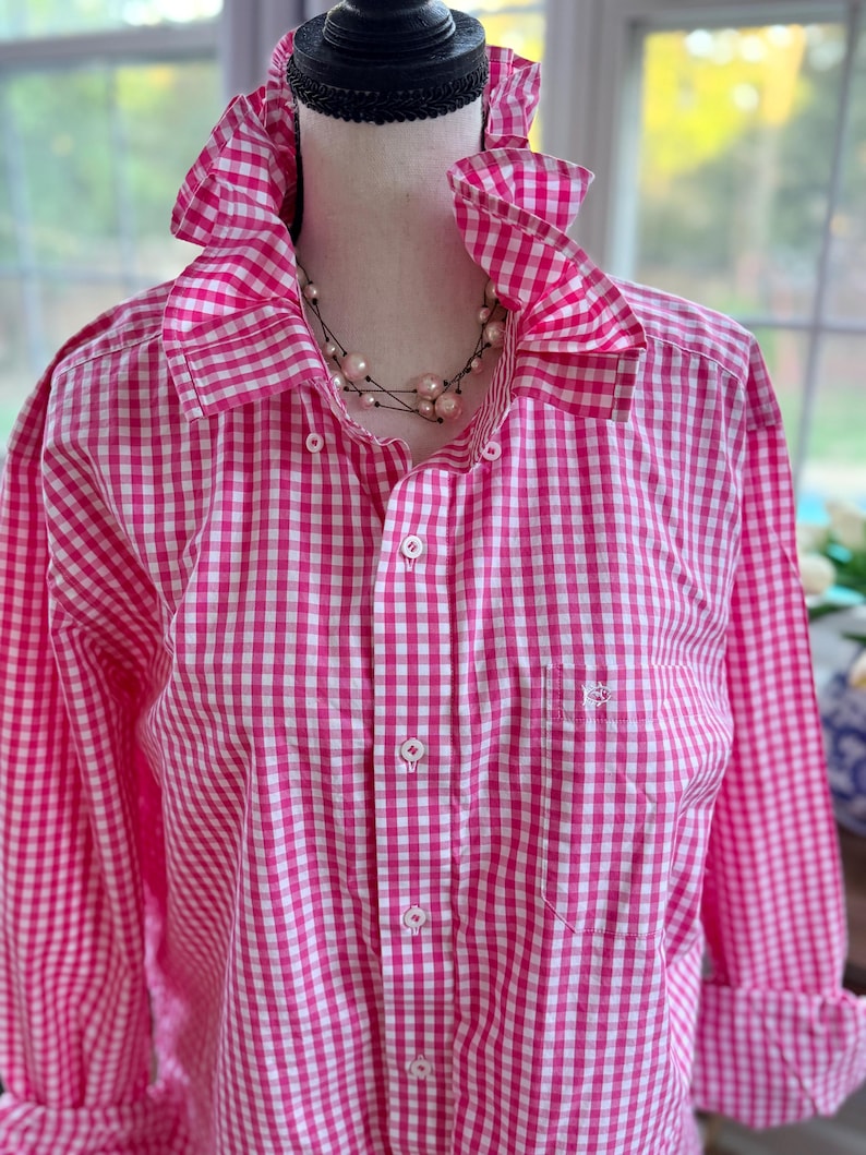 May include: A pink and white gingham button-down shirt with a ruffled collar and cuffs. The long-sleeved shirt has a small pocket on the chest and is displayed on a mannequin. A pearl necklace is visible.