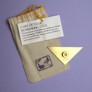 Puede incluir: Una herramienta de latón para hacer cajas de forma triangular con un remache circular, junto a una bolsa beige con cordón. Se adjunta una etiqueta con instrucciones de cuidado. El texto de la etiqueta dice "CARE OF YOUR BOXMAKING TOOL".