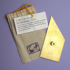 May include: A brass boxmaking tool with a geometric shape and a small, round knob. The tool is accompanied by a small, beige drawstring bag and a white tag with care instructions. The tag reads "CARE OF YOUR BOXMAKING TOOL".