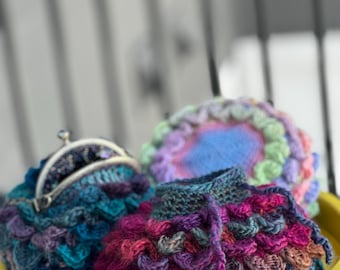 May include: Three crocheted coin purses in various shades of purple, blue, and green. The purses are sitting on a yellow table.