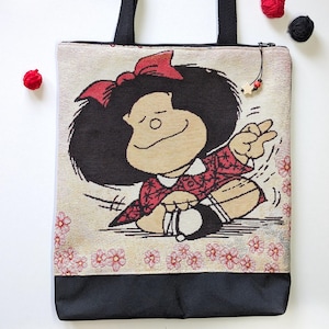 Puede incluir: Bolso tote con un personaje de dibujos animados con un gran afro negro y un lazo rojo. El bolso tiene asas negras y una base negra. El personaje viste un atuendo con estampado rojo y blanco. El fondo es beige con detalles florales rosas.