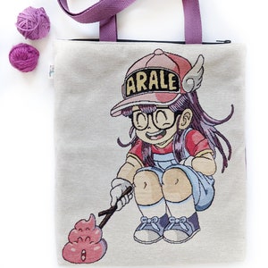Puede incluir: Una bolsa de lona blanca con cierre de cremallera y asas moradas. La bolsa presenta una imagen bordada de colores de una niña de dibujos animados con cabello rosado que lleva un sombrero rosado con la palabra "ARALE" en él. La niña sostiene un palo con un emoji de caca rosa en el extremo.