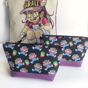 Puede incluir: Un conjunto de tres bolsas con una cremallera morada y un ribete morado. La bolsa más grande es una bolsa de mano con un frente de lona beige que presenta un personaje de dibujos animados con el texto "ARALE". Las dos bolsas más pequeñas son bolsas con cremallera con un fondo negro y un patrón repetido de un personaje de dibujos animados.