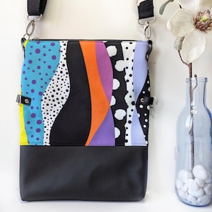 May include: A rectangular crossbody bag with a black leather bottom and a colorful patterned top. The top features vertical stripes in blue with purple dots, black, orange, purple, white with black dots, and light blue. The bag has a black adjustable strap.