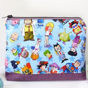 Puede incluir: Una bolsa azul con cremallera con un estampado colorido de personajes de dibujos animados. El estampado presenta una variedad de personajes, incluyendo un hombre con bigote, una mujer con el pelo largo y un extraterrestre verde. La bolsa tiene un forro de tela morado.