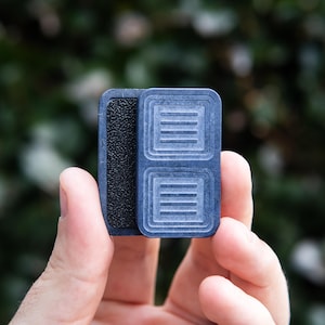 May include: A small, blue and black textured object with a rectangular shape. The object has a raised design with lines on the front.