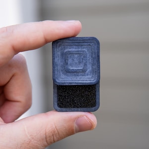 Puede incluir: Un objeto cuadrado negro con una superficie texturizada y un cuadrado más pequeño en el centro. El objeto se sostiene en una mano.
