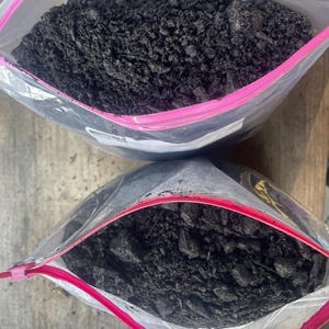 Puede incluir: Dos bolsas de plástico transparente con cierre de cremallera llenas de tierra oscura. Las bolsas tienen detalles rosas y rojos a lo largo de la cremallera. La tierra es de color marrón oscuro o negro, con una textura ligeramente áspera. Las bolsas están parcialmente abiertas.