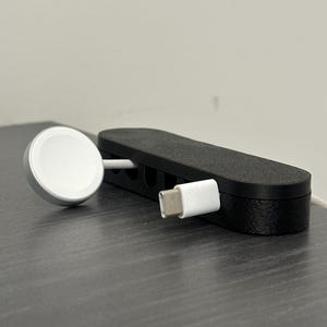 Puede incluir: Una base de carga negra para un Apple Watch y un cable USB-C. La base es rectangular con una superficie texturizada y varias ranuras. El cargador del reloj es blanco y plateado.
