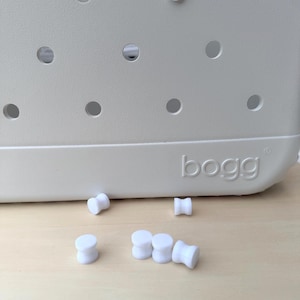 Puede incluir: Un bolso tote beige claro con un diseño perforado y el nombre de la marca "bogg" en relieve en el lateral. Varios tapones cilíndricos blancos están esparcidos cerca de la bolsa, algunos insertados en los agujeros.
