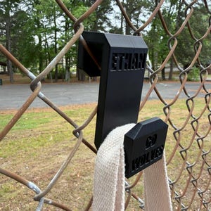 May include: Black 3D-printed fence hook with the name "ETHAN" and the text "GO WOLVES". A beige strap hangs from the hook, which is attached to a chain-link fence. The background features trees and grass.