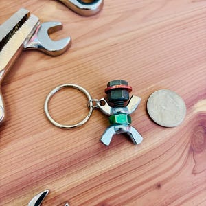 May include: A silver and green metal keychain shaped like a person made from nuts and bolts. The keychain is attached to a silver key ring.