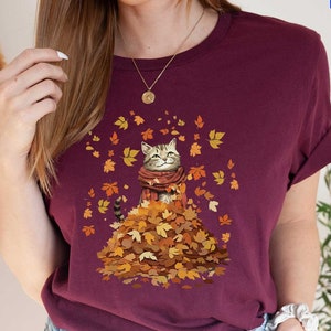 May include: A maroon t-shirt with a graphic of a cat sitting in a pile of autumn leaves. The cat is wearing a scarf and has a happy expression.