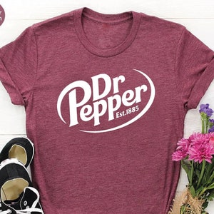 May include: Heather maroon t-shirt with the Dr Pepper logo in white. The shirt is paired with black canvas sneakers and a bouquet of pink and purple flowers. The text "Dr Pepper Est. 1885" is printed on the shirt.