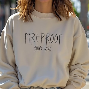 May include: A beige sweatshirt with the text "FIREPROOF since 1692" in black. The sweatshirt is a crew neck style. The model is smiling and wearing blue jeans.