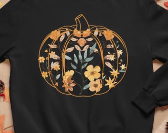 May include: Black sweatshirt featuring a floral pumpkin design. The pumpkin is outlined in gold and filled with yellow flowers and green leaves. The sweatshirt is surrounded by fall foliage, pumpkins, and pinecones.