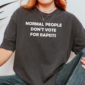 May include: Dark grey crew neck t-shirt featuring the white text: "NORMAL PEOPLE DON'T VOTE FOR RAPISTS". The shirt is made of a soft material. The image shows the shirt being worn. The person is wearing a gold necklace.