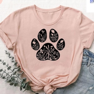 May include: A heather peach t-shirt featuring a black paw print design filled with floral and butterfly silhouettes. The shirt has a crew neck and short sleeves. The text "heather peach" is in a circle on the upper left.