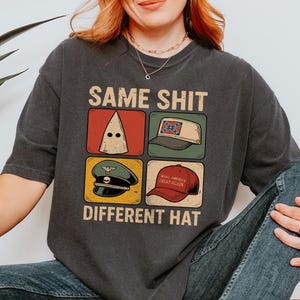 May include: Dark gray t-shirt with the text "SAME SHIT DIFFERENT HAT" above four images of hats. The hats include a white hood, a tan baseball cap with a Confederate flag, a green military hat, and a red "Make America Great Again" hat.