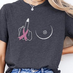 May include: Dark gray t-shirt with a graphic of a hand giving the middle finger, a pink ribbon, and a breast. The shirt is made of a soft material and is a crew neck style.