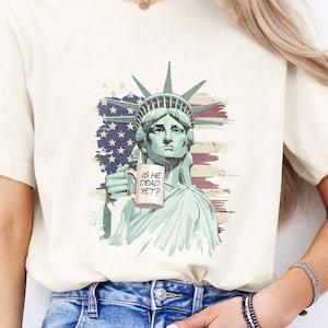 K&ouml;nnte beinhalten: Cremefarbenes T-Shirt mit einer Grafik der Freiheitsstatue, die ein Schild mit der Aufschrift "IS HE DEAD YET?" h&auml;lt. Das Design beinhaltet einen Hintergrund der amerikanischen Flagge. Das Shirt wird mit blauen Jeans getragen.