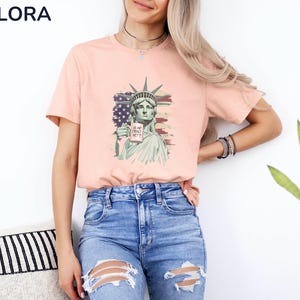 K&ouml;nnte beinhalten: Pfirsichfarbenes T-Shirt mit einer Grafik der Freiheitsstatue, die ein Schild mit der Aufschrift "IS SHE DEAD YET?" h&auml;lt. Das Design beinhaltet die amerikanische Flagge. Das T-Shirt hat einen Rundhalsausschnitt.