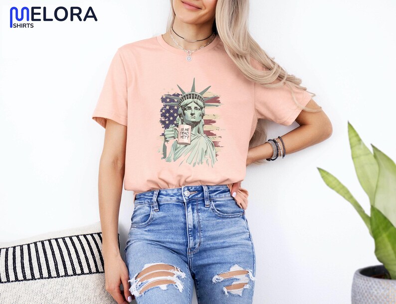 K&ouml;nnte beinhalten: Pfirsichfarbenes T-Shirt mit einer Grafik der Freiheitsstatue, die ein Schild mit der Aufschrift "IS SHE DEAD YET?" h&auml;lt. Das Design beinhaltet die amerikanische Flagge. Das T-Shirt hat einen Rundhalsausschnitt.