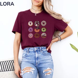 Puede incluir: Camiseta burdeos con una cuadr&iacute;cula de nueve ilustraciones de donuts con tem&aacute;tica de Halloween. Los donuts tienen varios ingredientes y decoraciones, incluyendo un fantasma, un sombrero de bruja y una calavera. La camiseta tiene el texto "MELORA SHIRTS" en la parte superior izquierda.