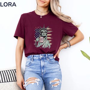 K&ouml;nnte beinhalten: Burgunderfarbenes T-Shirt mit einer Grafik der Freiheitsstatue, die eine Tasse mit dem wei&szlig;en Text "IS HE DEAD YET?" h&auml;lt. Die Grafik enth&auml;lt ein amerikanisches Flaggen-Design. Das Shirt wird mit blauen Jeans getragen.