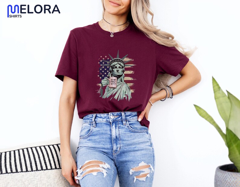 K&ouml;nnte beinhalten: Burgunderfarbenes T-Shirt mit einer Grafik der Freiheitsstatue, die eine Tasse mit dem wei&szlig;en Text "IS HE DEAD YET?" h&auml;lt. Die Grafik enth&auml;lt ein amerikanisches Flaggen-Design. Das Shirt wird mit blauen Jeans getragen.
