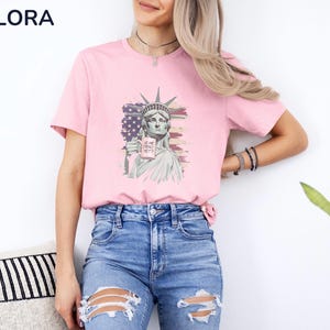 K&ouml;nnte beinhalten: Hellrosa T-Shirt mit einer Grafik der Freiheitsstatue, die ein Schild mit der Aufschrift "IS HE DEAD YET?" h&auml;lt. Die Grafik ist vor dem Hintergrund der amerikanischen Flagge platziert. Das Shirt wird mit blauen Jeans getragen.