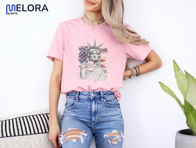 K&ouml;nnte beinhalten: Hellrosa T-Shirt mit einer Grafik der Freiheitsstatue, die ein Schild mit der Aufschrift "IS HE DEAD YET?" h&auml;lt. Die Grafik ist vor dem Hintergrund der amerikanischen Flagge platziert. Das Shirt wird mit blauen Jeans getragen.