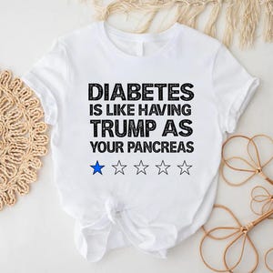 Puede incluir: Camiseta blanca con texto negro que dice "La diabetes es como tener a Trump como p&aacute;ncreas". Hay cinco estrellas negras y una estrella azul debajo del texto.
