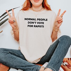 May include: Cream-coloured t-shirt with the text "NORMAL PEOPLE DON'T VOTE FOR RAPISTS" in black. The shirt is worn by a person wearing blue jeans and making a peace sign with their fingers.