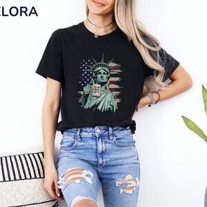 K&ouml;nnte beinhalten: Schwarzes T-Shirt mit einer Grafik der Freiheitsstatue, die ein Schild mit der Aufschrift "IS SHE DEAD YET?" h&auml;lt. Das Design beinhaltet die amerikanische Flagge. Das T-Shirt hat einen Rundhalsausschnitt.