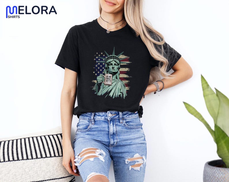 K&ouml;nnte beinhalten: Schwarzes T-Shirt mit einer Grafik der Freiheitsstatue, die ein Schild mit der Aufschrift "IS SHE DEAD YET?" h&auml;lt. Das Design beinhaltet die amerikanische Flagge. Das T-Shirt hat einen Rundhalsausschnitt.