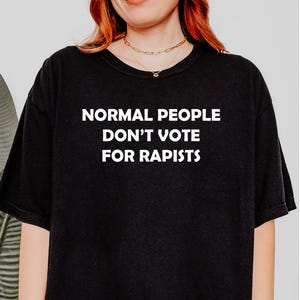 May include: Black t-shirt with the white text "NORMAL PEOPLE DON'T VOTE FOR RAPISTS". The shirt is a crew neck style.