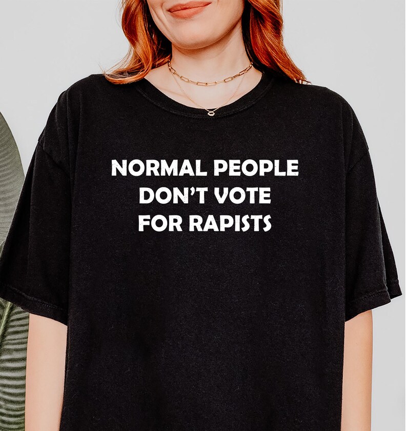 May include: Black t-shirt with the white text "NORMAL PEOPLE DON'T VOTE FOR RAPISTS". The shirt is a crew neck style.