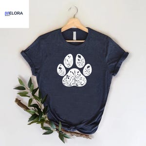 May include: Navy blue t-shirt featuring a white paw print design filled with floral silhouettes. The shirt is displayed on a wooden hanger. The brand name "MELORA SHIRTS" is visible in the upper left corner.