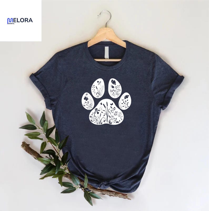 May include: Navy blue t-shirt featuring a white paw print design filled with floral silhouettes. The shirt is displayed on a wooden hanger. The brand name "MELORA SHIRTS" is visible in the upper left corner.