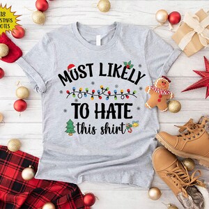Może przedstawiać: Szary świąteczny T-shirt z napisem "Most Likely To Hate This Shirt" i łańcuchem światełek choinkowych. Koszulka ma świąteczny wz&oacute;r z małym drzewem choinkowym i dzwonkiem.